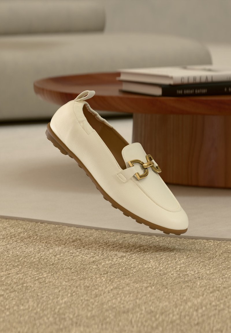 Μπεζ δερμάτινο loafer με χρυσή αγκράφα και λαστιχένια σόλα, τοποθετημένο σε υφασμένο μπεζ χαλί, με ξύλινο τραπεζάκι καφέ και βιβλία στο φόντο.