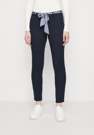 Laivastonsiniset slim-fit-housut, joissa sinivalkokuvioitu kangasvyö solmittuna vyötärölle, yhdistettynä valkoisiin tennareihin ja valkoiseen pitkähihainen puseroon.