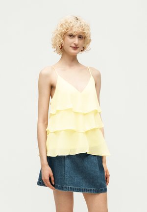 Personne aux cheveux blonds bouclés portant un haut jaune sans manches à volants et une mini-jupe en jean bleue, debout devant un fond uni.