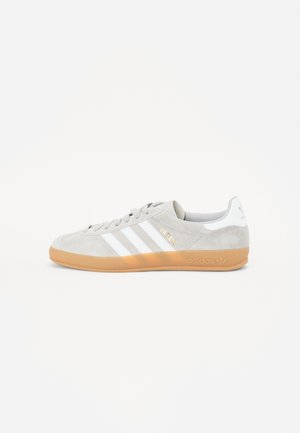 Baskets Adidas Gazelle grises avec des bandes blanches et une semelle en gomme, vue de côté sur fond blanc uni.