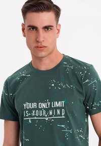 Mørkegrøn bomulds t-shirt med rund hals, prydet med en hvid tekstprint og teal sprøjtetilbehør over hele stoffet.