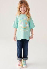 Mintgrøn t-shirt med farverigt grafisk design af et smilende ansigt og regnbue, parret med mørkeblå denimjeans og multicolore sneakers.