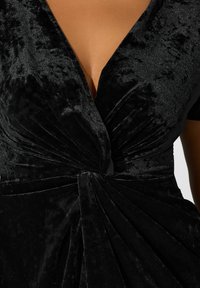 Mustassa samettimekossa on syvä V-pääntie, lyhyet hihat sekä kerrostettu, kierretty muotoilu vyötäröllä, jossa on teksturoitu pinta.