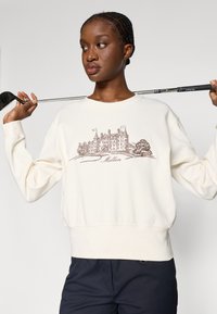 Sudadera de color crema con un corte relajado, que presenta una ilustración impresa en marrón de una mansión y el texto "Malbon" debajo.