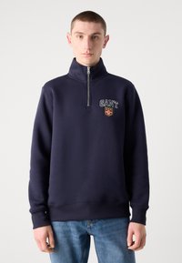 Tricou bleumarin din bumbac cu guler înalt și design cu fermoar pe jumătate. Dispune de logo-ul GANT și un patch cu scut. Material moale din bumbac, cu manșete ribbate.