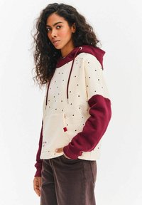 Bluza z kapturem z długimi rękawami, kremowym tułowiem w czarne groszki, bordowymi rękawami, ściągaczami i przednią kieszenią. Wykonana z miękkiego materiału.