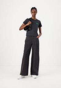 Svart avkortet t-skjorte med grønn logo, paret med løstsittende kolgrå bukser og hvite joggesko. Modellen holder en liten svart håndveske.