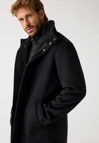 Cappotto nero in misto lana con colletto alto, strato interno con zip e bottoni a pressione. Presenta un design elegante e tasche laterali. Tessuto morbido.