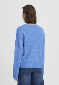 Pull en maille bleu à coupe décontractée, ourlet et poignets côtelés, texture douce et encolure ronde large. Porté avec un jean denim foncé.