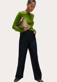 Grüner Strickpullover, lockere weite Jeans aus dunklem Denim und eine strukturierte beige Clutch. Flache Schuhe runden das Outfit ab. Einfaches und modernes Design.