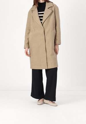 Femme portant un manteau texturé beige mi-long, un pantalon noir à jambes larges, un haut rayé noir et blanc, et des chaussures plates beiges.