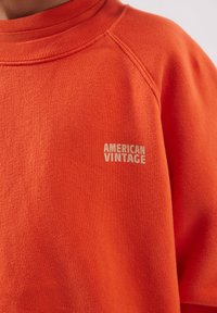 Makro snimka narančaste majice s rebrastim ovratnikom i natpisom "AMERICAN VINTAGE" svijetle bež boje na lijevoj strani prsa.