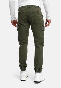 Homme portant un pantalon cargo vert olive et des baskets blanches, vu de dos, debout devant un fond uni.