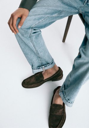 Person siddende på en stol iført lyseblå jeans, en mørk langærmet top og brune ruskindssko uden snørebånd, med den ene hånd hvilende på knæet.