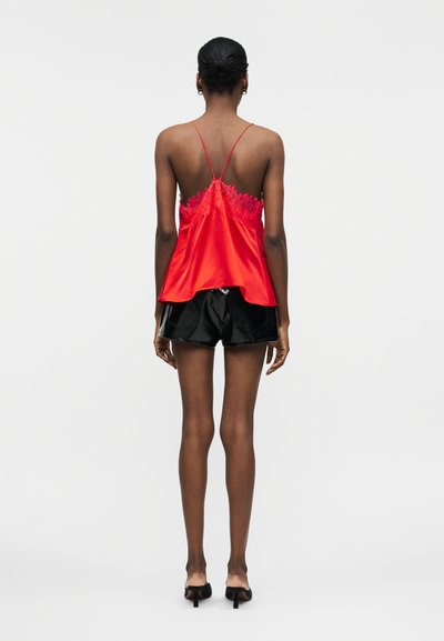 Γυναίκα με σκουρόχρωμο δέρμα που φοράει κόκκινο σατέν κορμάκι με δαντέλα, μαύρα γυαλιστερά σορτς και μαύρα mule με τακούνι, που στέκεται στραμμένη με την πλάτη προς τα εμπρός σε λευκό φόντο.