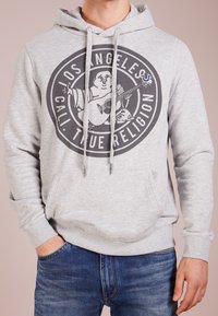 Sudadera gris con capucha y un gráfico redondo que presenta a un personaje tocando la guitarra, con cordones y detalle de bolsillo frontal. Se combina con unos vaqueros azules.