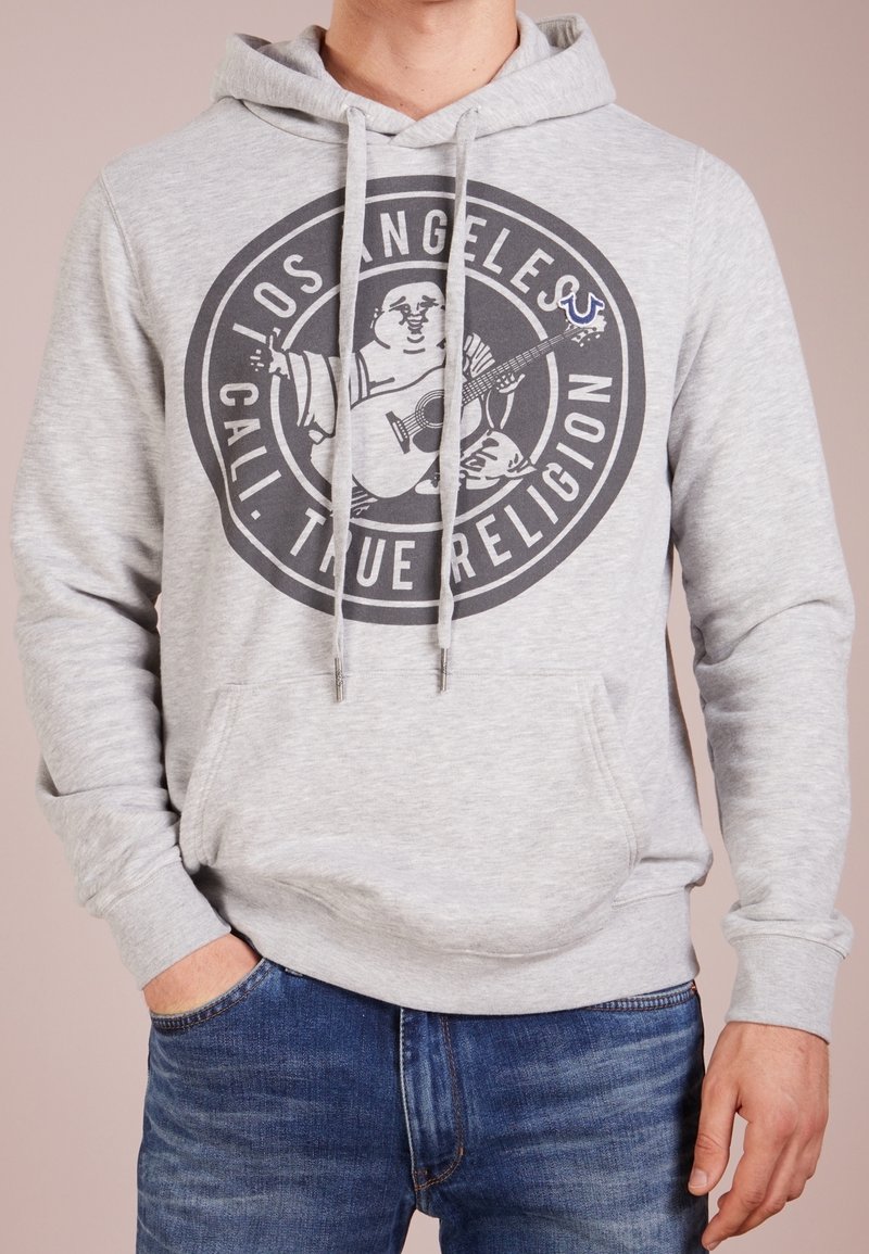 Sudadera gris con capucha y un gráfico redondo que presenta a un personaje tocando la guitarra, con cordones y detalle de bolsillo frontal. Se combina con unos vaqueros azules.