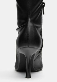 Fekete bőrből készült, magas sarkú csizma, amely illeszkedő szabással, hegyes orral és oldalsó cipzárral rendelkezik. A sarok stiletto stílusú és karcsú. Simább textúra.