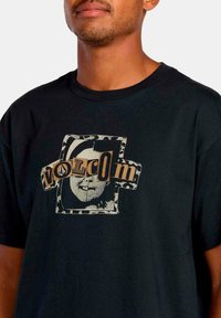 Camiseta de algodón negra con un gráfico destacado que presenta la palabra "Volcom" y un diseño de cara en beige y blanco, con un fondo texturizado.