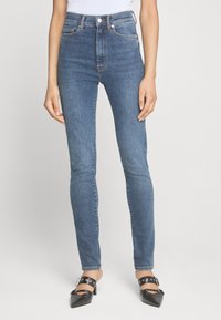 Jean skinny bleu avec taille haute, fabriqué en denim. Présente un zip à boutons, cinq poches et un délavage subtil sur les jambes.