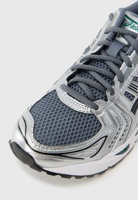 Γκρι αθλητικό παπούτσι με επάνω μέρος από δίχτυ και ασημί λεπτομέρειες, με στρογγυλεμένο δάκτυλο, σύστημα κορδονιών και υφασμένο ελαστικό πάτο για πρόσφυση.