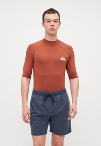 Rash guard i rust orange, kortermet, med en tettsittende design og en hvit logo. Kombinert med blå badebukser med elastisk midje.