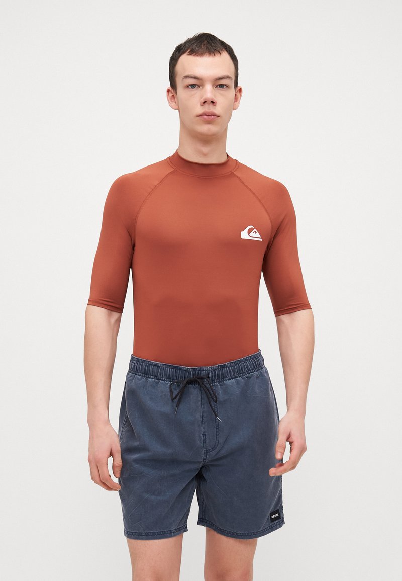 Rash guard i rust orange, kortermet, med en tettsittende design og en hvit logo. Kombinert med blå badebukser med elastisk midje.