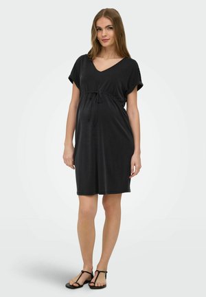 Žena v čiernych šatách po kolená s krátkymi rukávmi a so sťahovacím pásom v páse, stojaca a nosiaca čierne sandále na jednofarebnom pozadí.