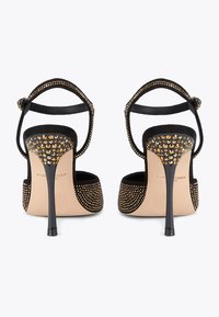 Sandali neri con tacco alto e cinturini alla caviglia, decorati con borchie dorate sui tacchi e sui cinturini, mostrati da dietro su uno sfondo bianco.