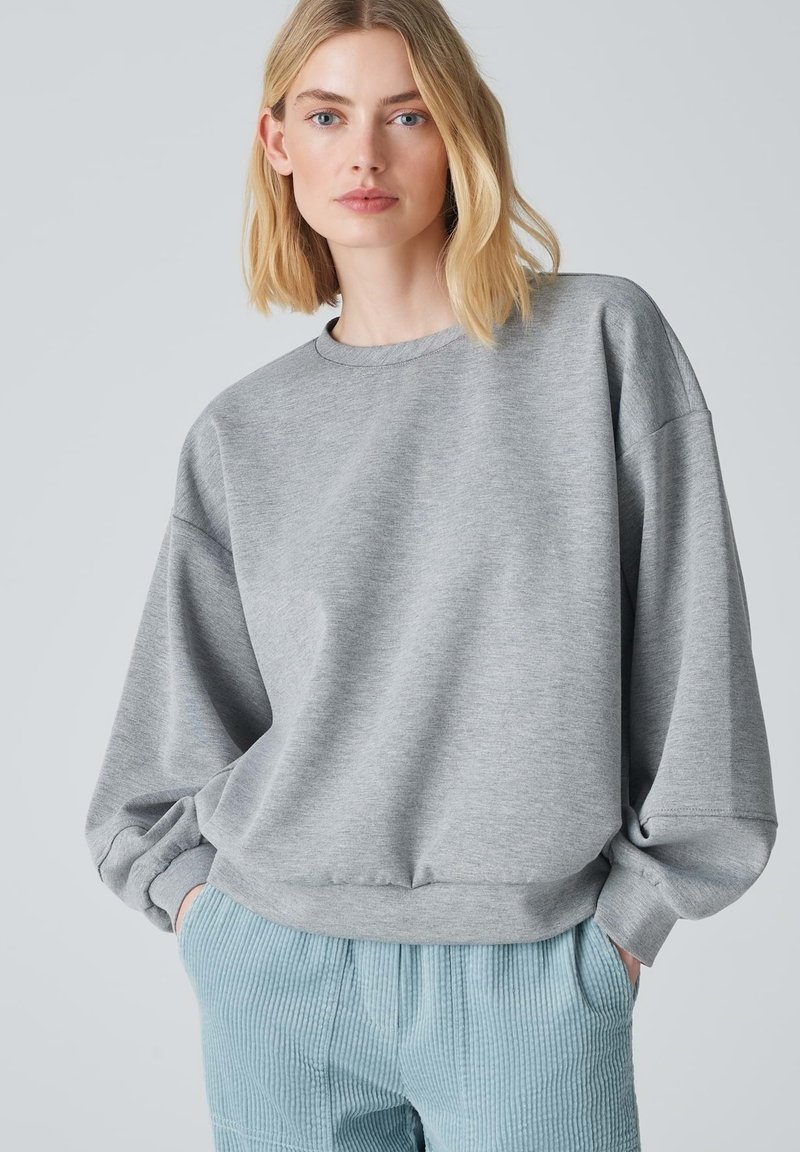 Felpa grigia oversized con spalle scese, polsini a costine e scollatura rotonda. Abbinata a pantaloni in corduroy blu chiaro.
