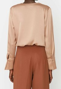 Langærmet beige satinbluse med rund halsudskæring og manchetter, parret med brune plisserede bukser. Glat tekstur og afslappet pasform.