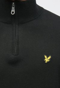 Fekete cipzáras galléros pulóver bordázott mintával, szoros gallérral és sárga hímzett sas logóval a bal mellkason.