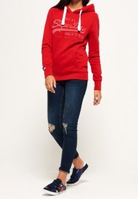 Rode capuchontrui met witte trekkoorden en logo, gecombineerd met donkerblauwe skinny jeans met versleten knieën en marineblauwe sneakers.