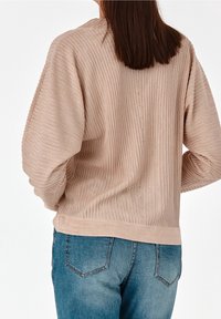 Pull beige clair à côtes avec une coupe décontractée, doté d'un col rond et de détails subtils le long de la couture arrière. Associé à un jean bleu.