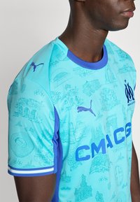 Ανοιχτό μπλε αθλητική φανέλα με μπλε λεπτομέρειες, που διαθέτει υφή με μοτίβο τοποθεσιών, το λογότυπο της Puma και έντονη επιγραφή «CMAG» στο μπροστινό μέρος.