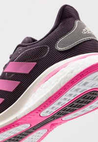 Sportovní obuv s fialovým textilním svrškem, růžovými pruhy a bílou mezipodešví. Výrazný šedý akcent na patě a texturovaný vzor podešve.