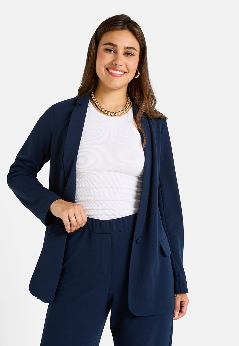 Blazer ajusté bleu marine en matériau doux, présentant un bouton unique, des revers en notch et deux poches avant, associé à un top blanc à côtes.