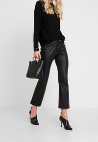 Schwarzer Strickpullover mit Nieten-Halsausschnitt, kombiniert mit schwarzen Lederhosen und schwarzen Slingback-High Heels. Kleine schwarze Handtasche mit goldenen Akzenten.