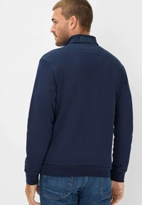 Navy blue sweatshirt with high collar, ribbed cuffs, and hem. Features subtle branding on the collar. Worn with light blue jeans.