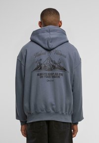 Grå hettegenser med en stor rygggrafikk av fjell og tekst. Løs passform med ribbede mansjetter og hem. Myk tekstur.
