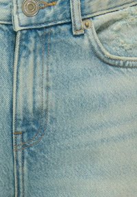 Gaiši zili džinsi ar izbalējušu tekstūru, redzamām šuvēm kontrastējošā brūnā krāsā un ar sudraba pogu akcentu jostasvietā.