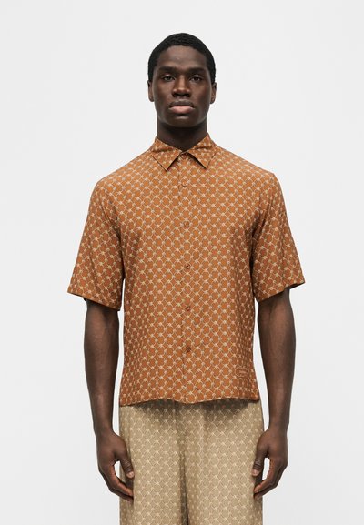 Άνδρας που φοράει καφέ πουκάμισο με κοντά μανίκια και λευκό γεωμετρικό σχέδιο, καθώς και μπεζ σορτς με σχέδιο, στέκεται μπροστά σε απλό φόντο.