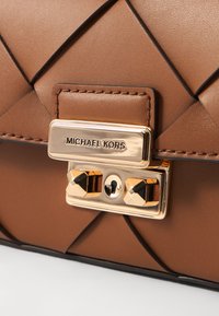 Borsa a mano in pelle marrone con design trapuntato, attrezzatura dorata e una placca logo prominente. Dotata di chiusura a scatto e borchie decorative.