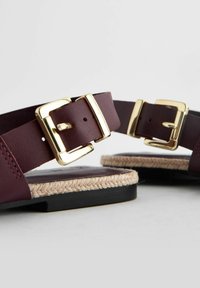 Sandales bordeaux avec deux boucles dorées ; fabriquées en cuir avec une bordure en jute tissé et une semelle en caoutchouc noir.