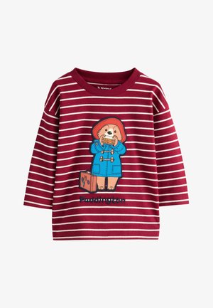 Cămașă cu mânecă lungă pentru copii, în dungi roșii și albe, cu Paddington Bear îmbrăcat într-un palton albastru, mâncând un sandwich, cu o valiză alături.