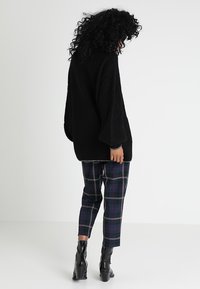 Przesadnie duży czarny sweter dzianinowy z teksturowanym wzorem, zestawiony z spodniami w kratę w kolorach granatowym, zielonym i bordowym oraz czarnymi botkami za kostkę.