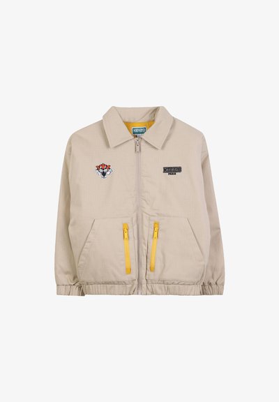 Veste zippée beige avec des poignets élastiques, deux poches zippées jaunes, logo Kenzo Paris sur la poitrine droite et patch de tête de tigre sur la poitrine gauche.