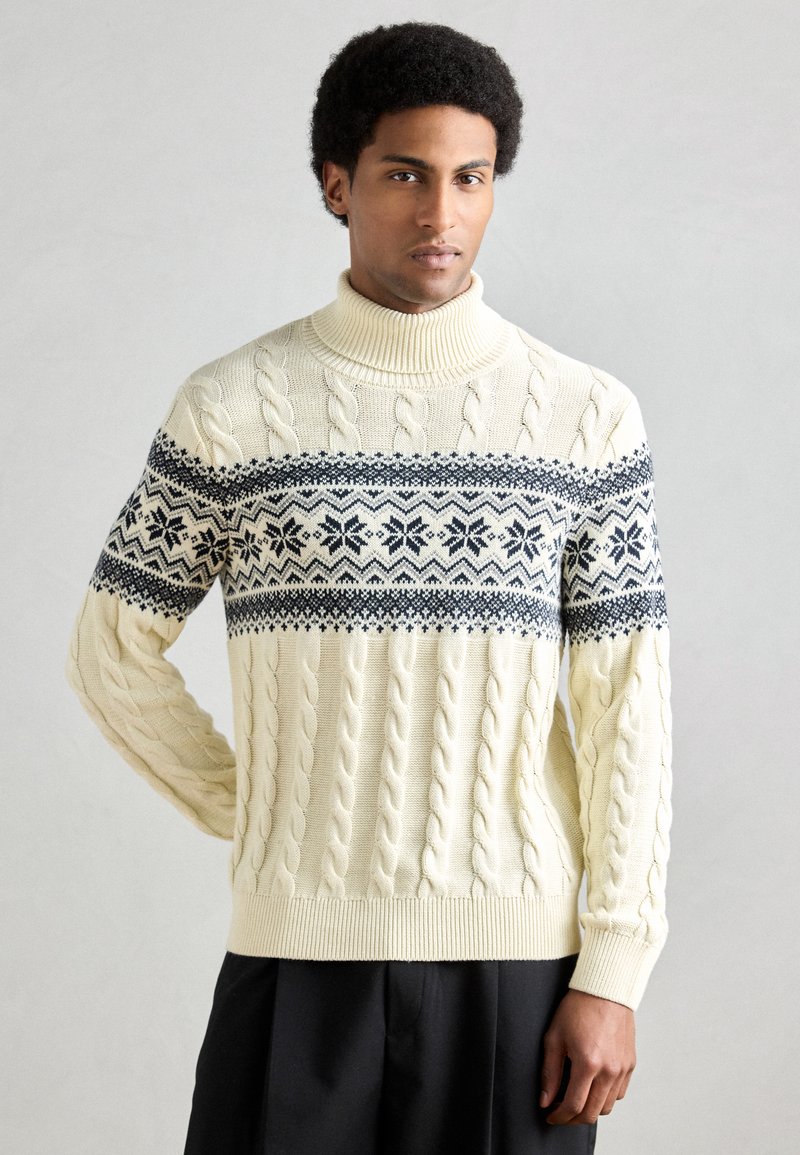 Camisola de malha cor de creme com gola alta. Apresenta um padrão de trança em malha e um desenho geométrico azul-marinho no peito.