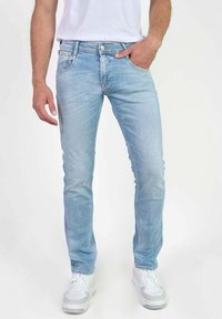 Jeans de mezclilla elásticos de color azul claro con corte ajustado, cinco bolsillos y cierre de botón. Llevados con una camiseta blanca y zapatillas blancas.