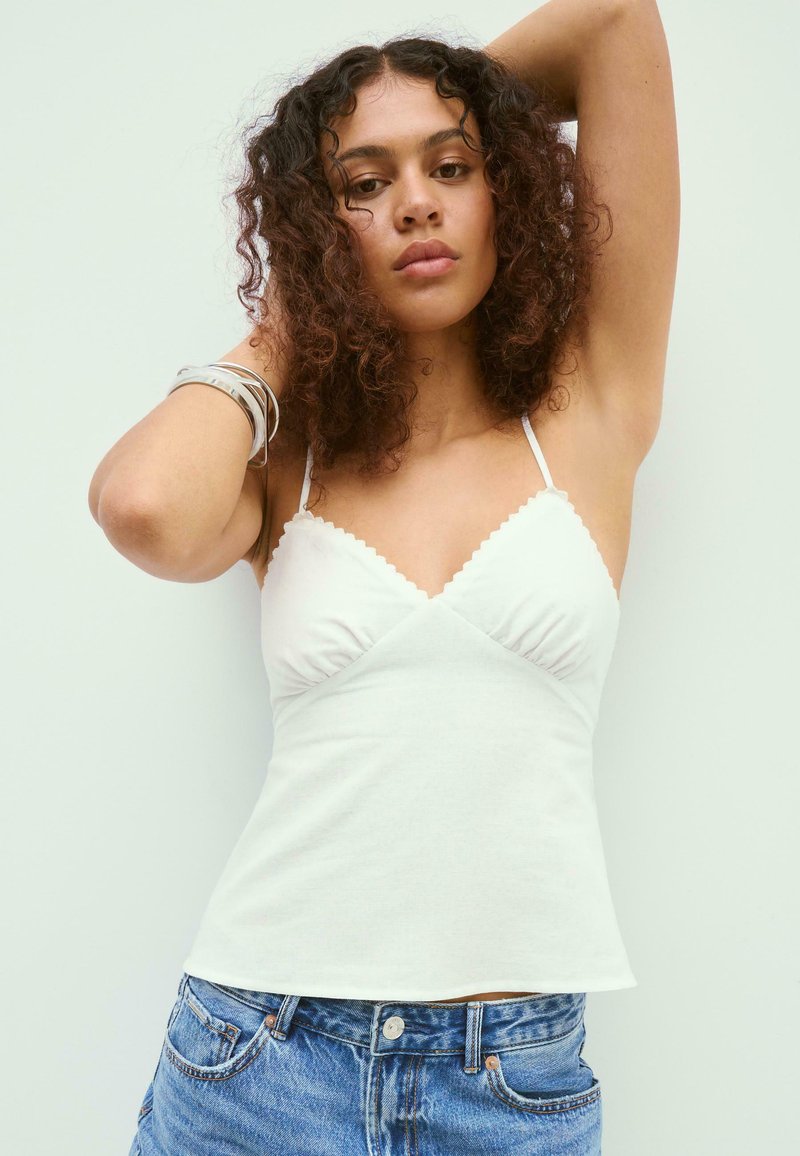 Femme aux cheveux bouclés portant un caraco blanc à bord festonné et un jean bleu, levant un bras et touchant ses cheveux de l'autre main.